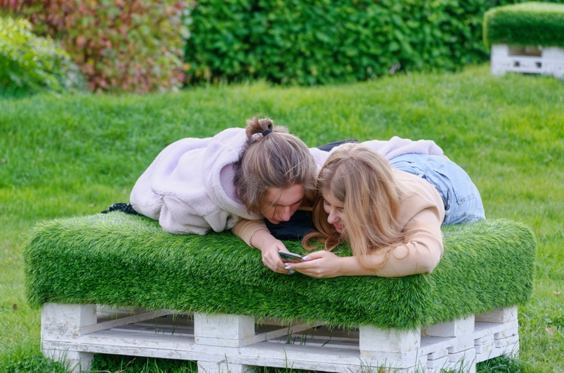 16 - Two girls watching a smartphone above a bench made from stacked wooden pallets covered with artificial green turf